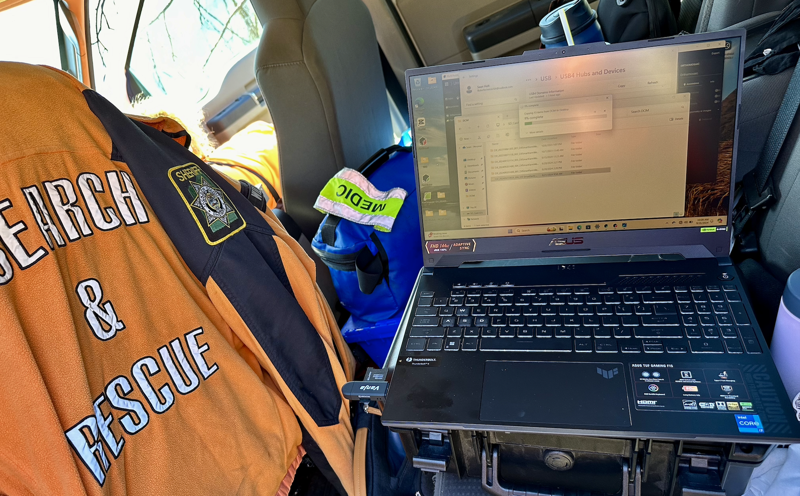 A laptop on a rugged case in a truck cab pulling drone flight data off a card, next to a Search and Rescue jacket with a sheriff patch and a medic bag