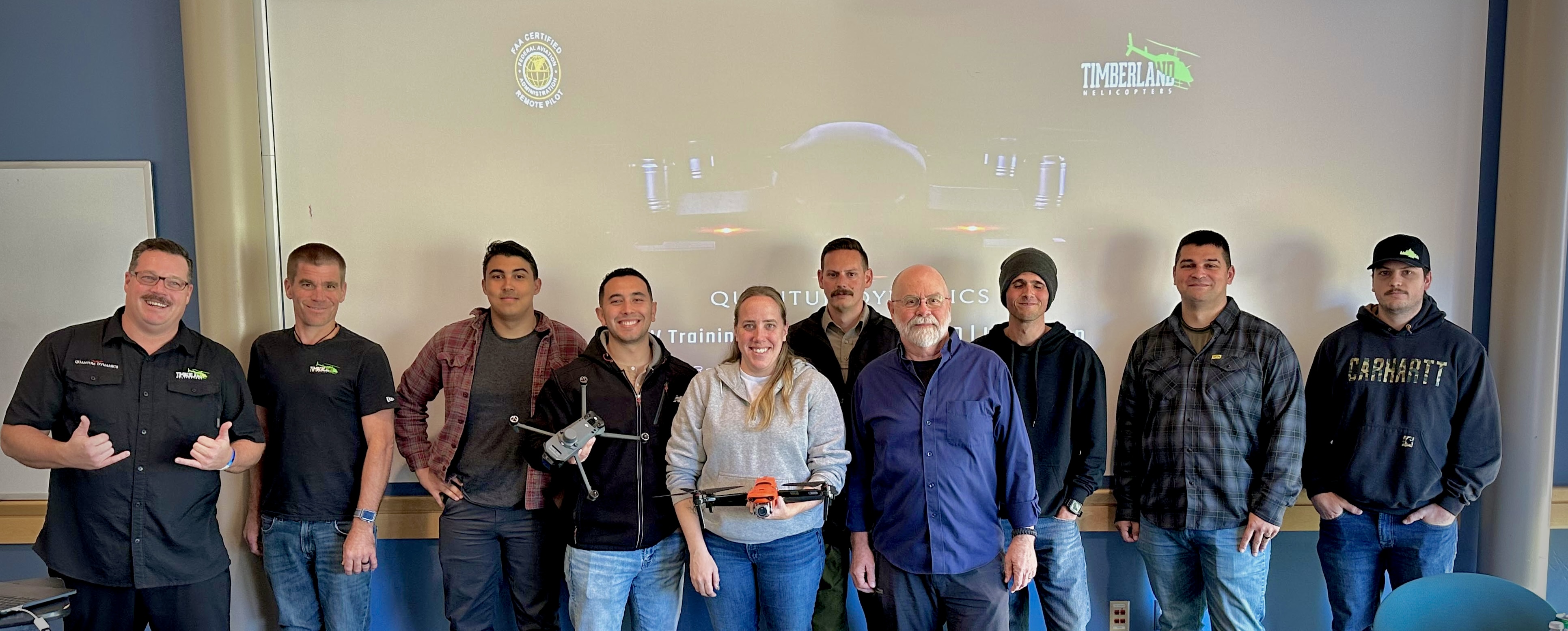 Task Force Almeda Class 002 group photo: ten students and instructors in a Quantum Dynamics classroom in front of the Timberland Helicopters training screen, two students holding drones